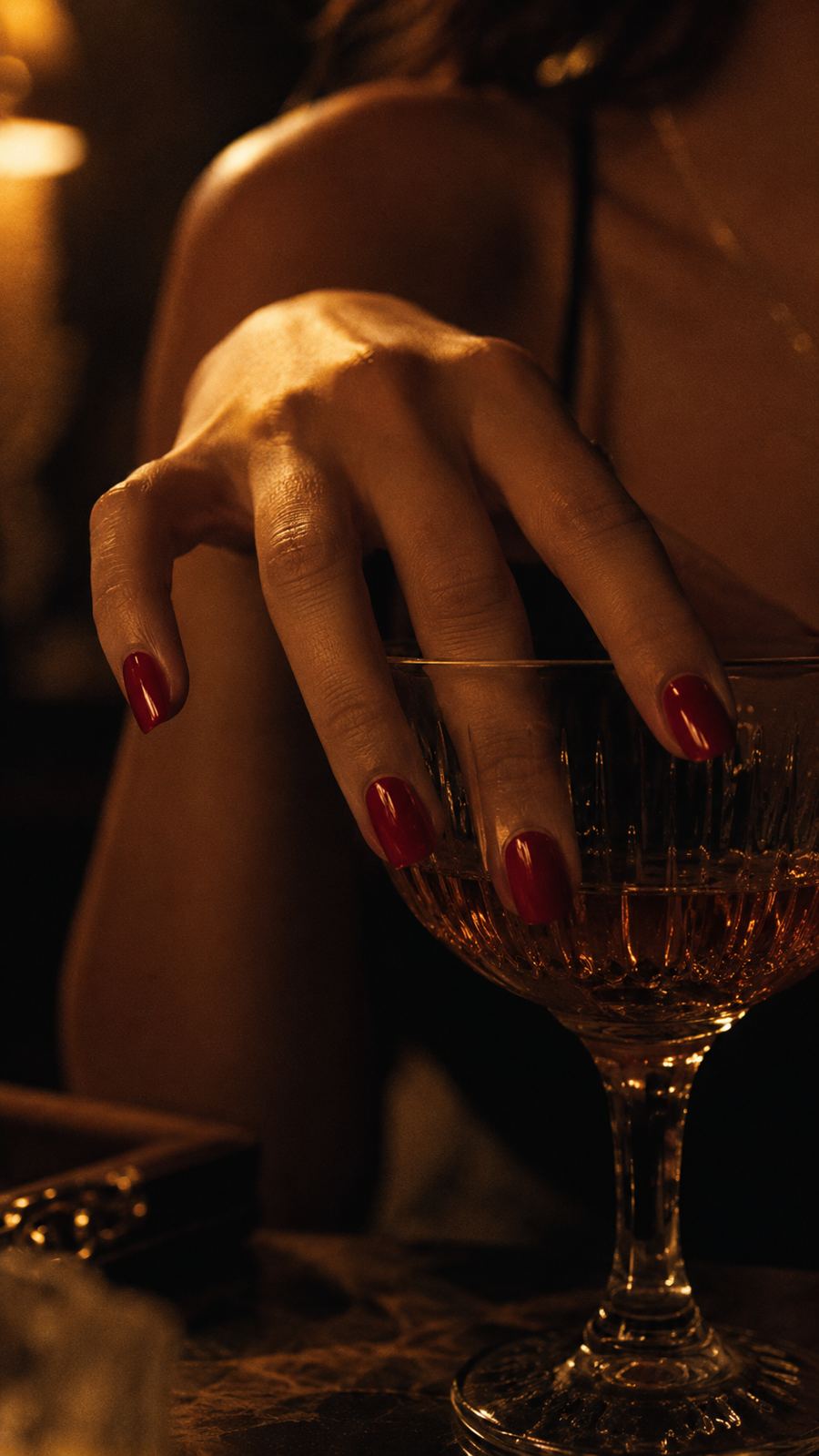 Une main aux ongles rouges sur le bord d’une coupe, lumière de bougie